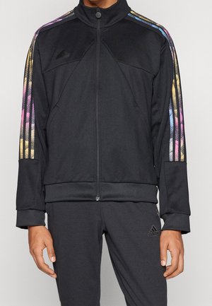 Homme portant un survêtement Adidas noir avec une fermeture éclair à l'avant et des bandes multicolores sur les manches et le pantalon.