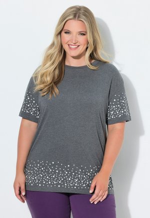 Femme souriante aux longs cheveux blonds portant un t-shirt gris à manches courtes avec des motifs de points blancs et un pantalon violet sur fond blanc.