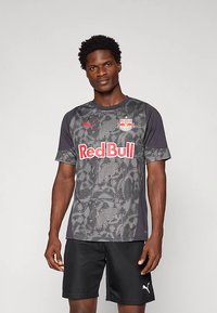 Harmaa urheilupaita, jossa naamiointikuvio, punainen "Red Bull" -logo, Puma-brändäys, lyhyet hihat ja pyöreä pääntie. Mustat shortsit.