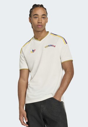 Joven con cabello trenzado que lleva una camisa blanca de manga corta con la palabra "Venezuela" y rayas de colores, posando con una mano en el bolsillo de sus pantalones vaqueros negros.