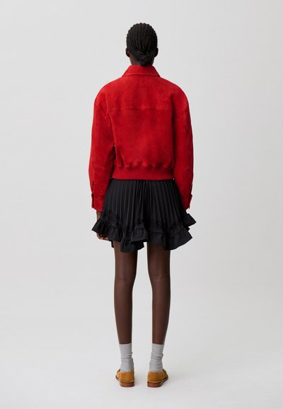 Giacca bomber in suede rosso con polsini a costine, abbinata a una gonna plissettata nera con orlo arricciato. Calzini grigi e scarpe marroni completano l'insieme.