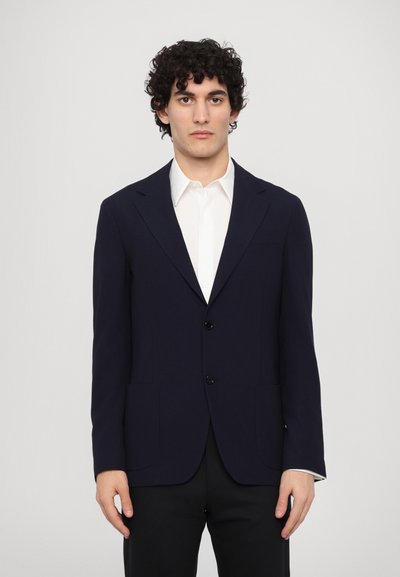 Blazer entallado azul marino con dos botones, solapas de muesca y bolsillos frontales. Lleva puesto sobre una camisa blanca, diseñado para una silueta ajustada.
