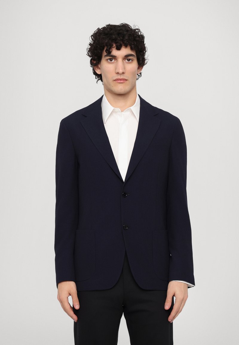 Blazer su misura blu navy con due bottoni, revers a taglio e tasche frontali. Indossato sopra una camicia bianca, progettato per una silhouette aderente.