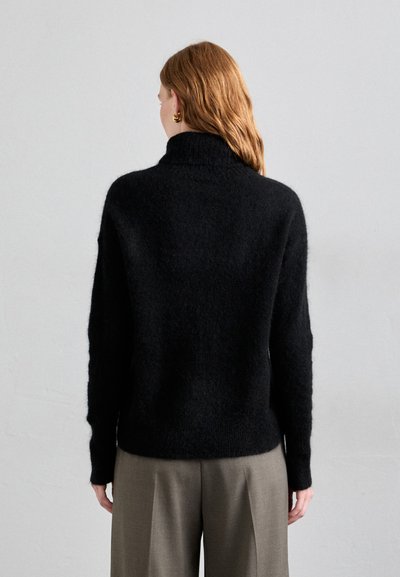 Black, soft, textured sweater with a high collar and relaxed fit. Worn with light-colored, wide-legged trousers.
