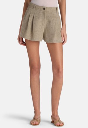 Kvinde iført beige plisserede hørshorts med knap- og lynlåslukning, kombineret med remsandaletter med flad sål på hvid baggrund.