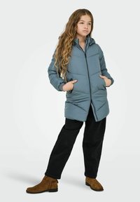 Puffer jacket in light blue with a zip closure, two side pockets, and a hood. Worn over black pants and brown suede ankle boots.