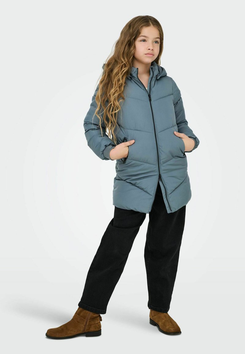 Puffer jacket in light blue with a zip closure, two side pockets, and a hood. Worn over black pants and brown suede ankle boots.