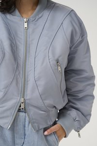 Giubbotto bomber di un grigio chiaro con design trapuntato, cerniere argentate, polsini a costine e due tasche laterali con cerniera. Finitura liscia e texture imbottita.