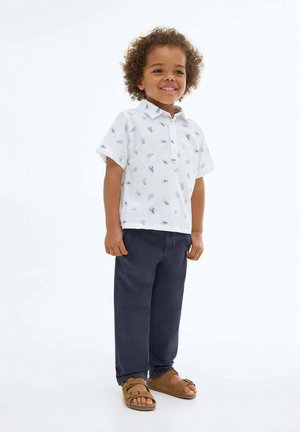 Enfant souriant avec des cheveux bouclés portant une chemise blanche à manches courtes avec des motifs de fourmis, un pantalon bleu marine et des sandales marron, se tenant sur un fond blanc.
