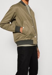 Oliivinvihreä pommitakki, jossa vetoketju edessä, resorit hihansuissa ja helmassa sekä kaksi sivutaskua. Sileä kangas ja metalliset nepparit.