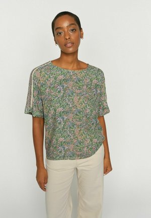 Frau mit kurzen Haaren, die ein grünes, kurzärmeliges Oberteil mit Blumenmuster und hellbeige Hosen trägt, steht vor einem schlichten hellen Hintergrund.