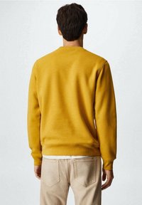 Gelbes Sweatshirt mit Rundhalsausschnitt, glatter Textur, gerippten Bündchen und Saum. Über hellen Hosen getragen, von hinten betrachtet.