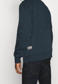 Sweatshirt G-Star bleu foncé avec un col rond, des poignets et un ourlet côtelés. Présente un patch logo en bas à gauche. Texture lisse.