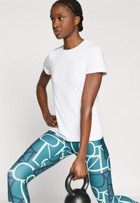 Λευκό αθλητικό κοντομάνικο πουκάμισο συνδυασμένο με τιρκουάζ κολάν με αφηρημένα γεωμετρικά σχέδια. Ο χρήστης κρατάει μια μαύρη αλτήρα kettlebell.