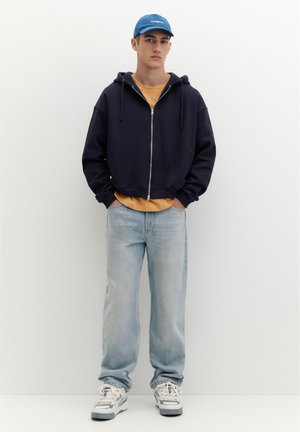 Jeune homme portant une casquette bleue, un sweat à capuche noir zippé, une chemise moutarde, un jean bleu clair et des baskets blanches, debout les mains dans les poches.