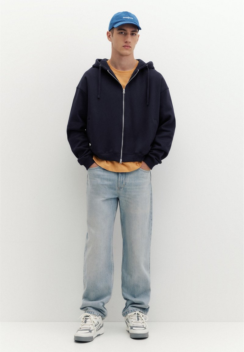 Jeune homme portant une casquette bleue, un sweat à capuche noir zippé, une chemise moutarde, un jean bleu clair et des baskets blanches, debout les mains dans les poches.