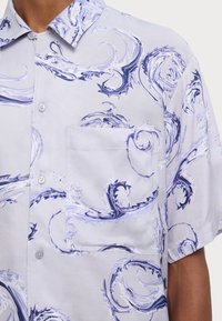 Camisa corta de manga corta en morado claro con patrones abstractos de remolinos en azul oscuro, bolsillo en la parte delantera y cuello abotonado, llevada por una persona.