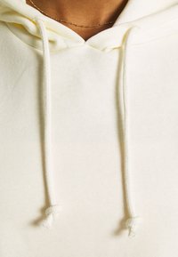 Sweat à capuche beige clair avec cordons ornés de nœuds, fabriqué en tissu doux et texturé ; mise au point sur la zone du cou et la capuche.