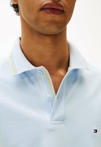 Polo shirt azzurro chiaro in tessuto texturizzato, con un accento giallo sul colletto e due bottoni. Logo visibile sulla patta.