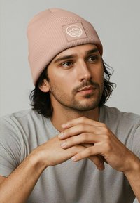 Pink ribbed knit beanie featuring a square patch with a mountain and wave design. Model wears a gray t-shirt, showing hands clasped.