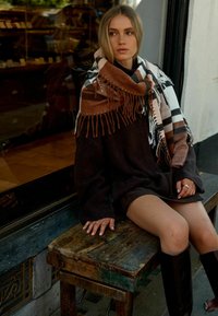 Overstørrelse mørk sweater, brun og hvid ternet tørklæde med frynser, kort nederdel, knæhøje brune støvler, siddende på en træstol.