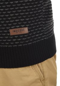 Mustvalge kirju kudum, millel on ribaga must äär, ja millel on pruun nahklabel, millel on kiri "INDICODE EST. 1993".