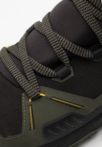 Scarpa sportiva nera con tessuto texturizzato, suola in gomma rinforzata, lacci intrecciati e accento giallo vicino al foro per i lacci.