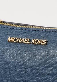 Blue textured fabric with a gold metallic "MICHAEL KORS" logo plaque and a visible gold zipper at the top edge.