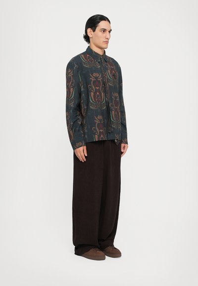 Button-up shirt in dark blue with intricate floral patterns in rust and gold, paired with wide brown trousers and brown sneakers.
