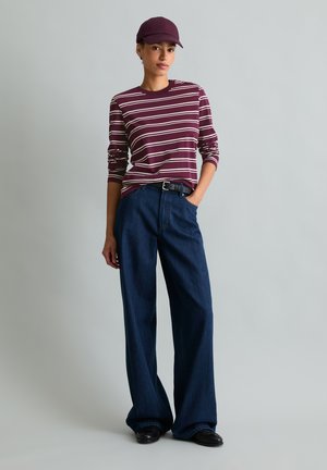 Jong persoon draagt een bordeauxrode gestreepte longsleeve, donkere wijdvallende jeans, zwarte schoenen en een bordeauxrode pet, staand met één hand in de zak.