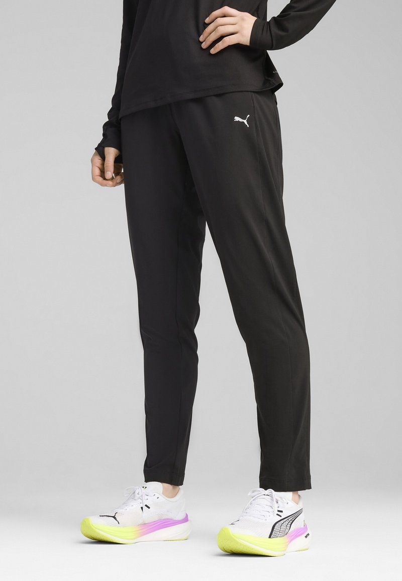 Zwarte sportieve broek met een slanke pasvorm, voorzien van een wit Puma-logo aan de linkerkant, gecombineerd met witte schoenen met een verloopzool in roze en groen.