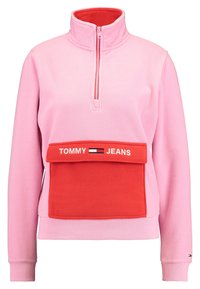 Polarowy sweter w jasnoróżowym kolorze z czerwonymi akcentami, z ćwierć zamkiem i dużą kieszenią z przodu, na której widnieje logo "TOMMY JEANS".