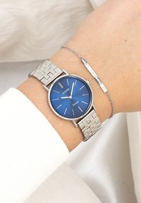 Montre en acier inoxydable argenté avec un cadran bleu, des aiguilles argentées et un bracelet à motif en grille. Accompagnée d'un bracelet fin en chaîne argentée.