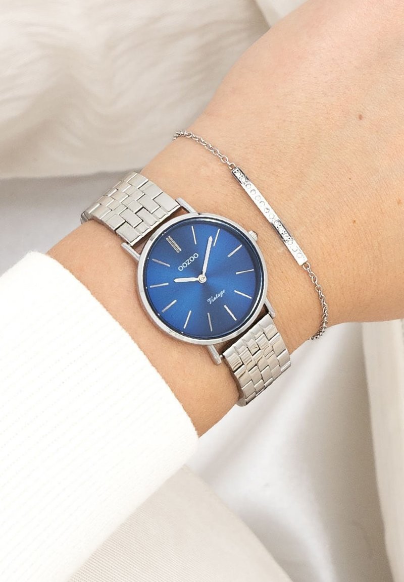 Montre en acier inoxydable argenté avec un cadran bleu, des aiguilles argentées et un bracelet à motif en grille. Accompagnée d'un bracelet fin en chaîne argentée.