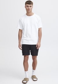 Weißes Baumwoll-T-Shirt mit kurzen Ärmeln, kombiniert mit schwarzen, strukturierten Shorts und beigen Slip-On-Sandalen, getragen mit weißen Sportsocken.