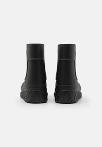 Botas de goma negras hasta el tobillo con un acabado texturizado, que cuentan con una punta redondeada y una suela gruesa con patrón. Lengüetas laterales dobles para facilitar su uso.