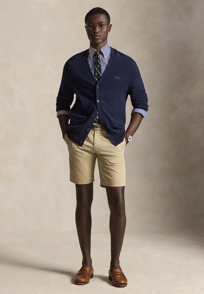 Mann iført marineblå cardigan over stripete skjorte og slips, beige shorts, brune loafers og runde briller, stående med hendene i lommene.