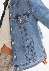 Veste en denim bleu clair avec fermeture à boutons, deux poches poitrine et poches latérales. Présente une texture délavée et des coutures contrastées.