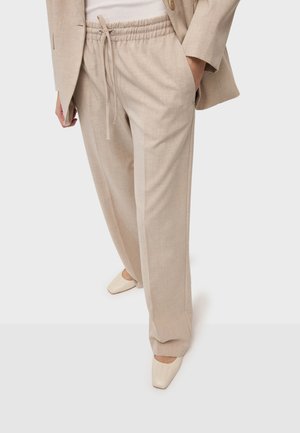 Kobieta ubrana w beżowe spodnie o szerokich nogawkach z troczkiem, pasujący blezer, biały top i kremowe buty z kwadratowym noskiem, stojąca z jedną ręką w kieszeni.