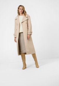 Beige dubbelknäppt kappa med stora slag, bärs över en krämfärgad stickad tröja och beige kjol. Kompletterad med ljust tanfärgade knähöga stövlar.