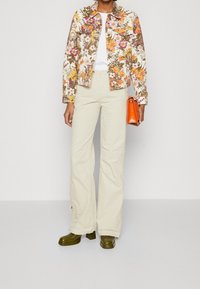 Giacca di jeans floreale bianca con fiori arancioni e viola vivaci; abbinata a pantaloni crema e scarpe col tacco verde scuro; porta una borsa arancione.