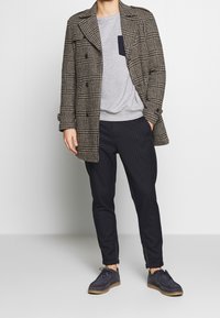 Abrigo de cuadros marrón con solapas anchas, sudadera gris con bolsillo en el pecho, pantalones de rayas azules y oscuros, y zapatos oscuros.