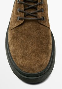 Chaussure en suede marron avec lacets noirs, semelle en caoutchouc et bout rond. Présente un dessus texturé et des coutures contrastantes.