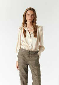 Blusa de satén crema con cuello tipo camisa y mangas abullonadas, combinada con pantalones de cuadros en tonos tierra, que cuentan con un cierre de botones.