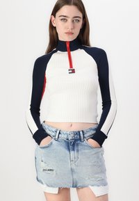 Fehér bordás, hosszú ujjú felső, tengerészkék és piros részletekkel, magas gallérral és fél cipzárral. Kopottas farmer mini szoknyával párosítva.