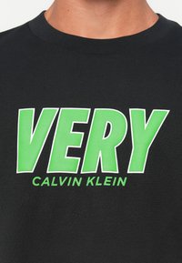 Muž v čiernom tričku s okrúhlym výstrihom, na ktorom je veľký zelený nápis "VEĽMI" a menší zelený nápis "CALVIN KLEIN" na hrudi.