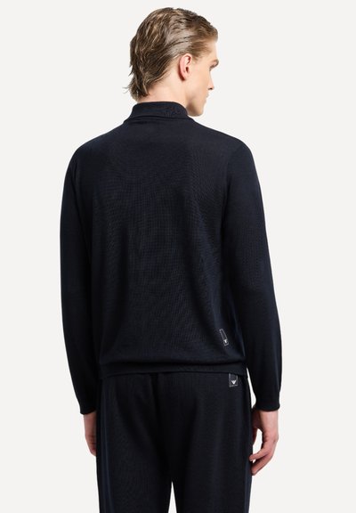 Maglione nero lavorato a maglia con collo alto, maniche lunghe e texture a coste. Presenta un dettaglio logo sul retro vicino all'orlo.