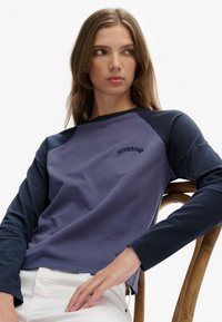 Jonge vrouw met lang bruin haar, gekleed in een marineblauwe en paarse lange mouwen shirt, zittend op een houten stoel en zijwaarts kijkend.