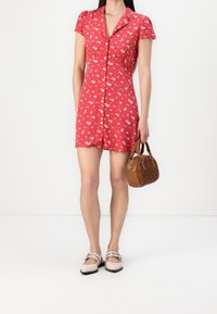 Rotes Kurzarmkleid mit floralem Muster, Knopfleiste und tailliertem Schnitt. Kombiniert mit einer braunen Handtasche und hellen Schuhen.