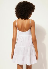 Weißes Baumwoll-Sommerkleid mit dünnen verstellbaren Trägern, gerafftem Oberteil und gestuftem Rüschensrock. Leichte Textur, lässiges Design.